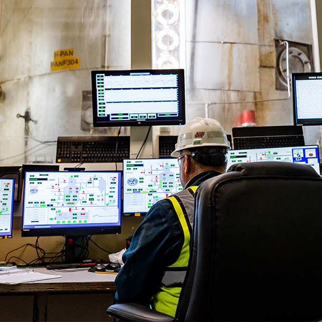 A U.S. Sugar operator in a hard hat and safety vest monitors multiple process control screens at a refinery control station, ensuring regulatory compliance and consistent product quality